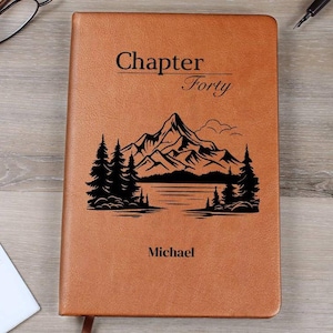 Peut inclure: Un journal en cuir marron avec les mots "Chapter Forty" et le nom "Michael" imprimés sur la couverture. Une illustration noire de montagnes, d'arbres et d'un lac est présentée sous le texte.