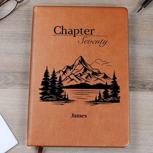 May include: A brown leather journal featuring the text "Chapter Seventy" and the name "James". The cover also displays a black mountain and lake scene.