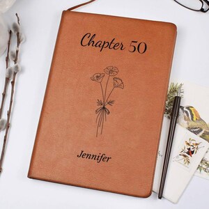 May include: A brown leather journal with the text "Chapter 50" and the name "Jennifer" engraved on the cover. A floral illustration is also featured. A black pen rests beside the journal.