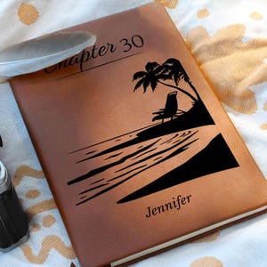 May include: A brown leather journal featuring the text "Chapter 30" and "Jennifer". The cover also has a black silhouette of a beach scene with a palm tree and a chair. A feather rests on the journal, which is on a patterned surface.