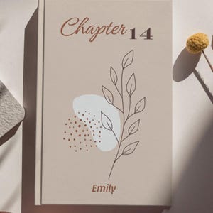 Può includere: Un libro con copertina rigida beige con il testo "Chapter 14" in corsivo marrone. Un'illustrazione botanica e il nome "Emily" sono anch'essi sulla copertina. Un fiore secco e un piccolo elemento decorativo sono sullo sfondo.