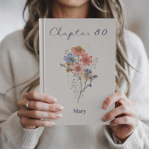 Puede incluir: Un libro de tapa dura con el texto "Chapter 80" en escritura cursiva y el nombre "Mary" impreso debajo de una ilustración floral. Las flores son en tonos rosa, naranja, azul y amarillo.