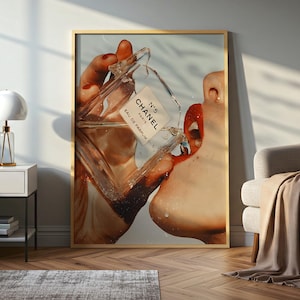 May include: Framed art print featuring a close-up of a person's face with red lips, drinking from a Chanel No. 5 perfume bottle. The bottle is clear with a white label. The artwork is in a gold frame.