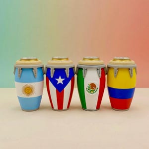 May include: Four cylindrical can coolers shaped like conga drums, each featuring a different national flag design: Argentina, Puerto Rico, Mexico, and Colombia. Each cooler has a tan top rim, a silver-colored band, and a tapered base design.