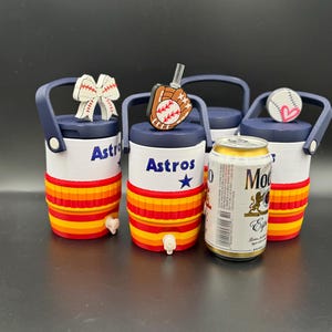 May include: Three small white and orange beverage coolers with navy blue lids and handles, each featuring the text Astros and a blue star. The lids are topped with baseball-themed charms including a bow, a glove, and a heart-accented baseball.