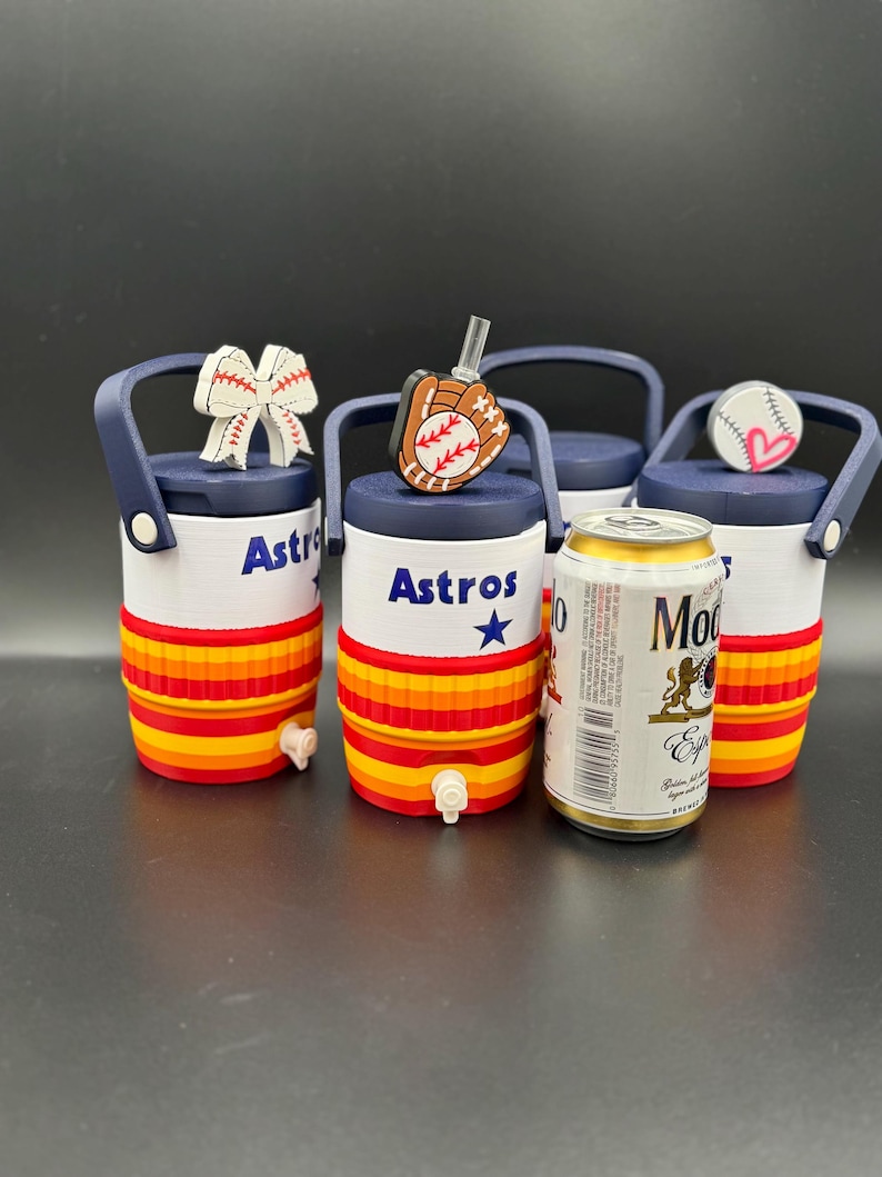 May include: Three small white and orange beverage coolers with navy blue lids and handles, each featuring the text Astros and a blue star. The lids are topped with baseball-themed charms including a bow, a glove, and a heart-accented baseball.