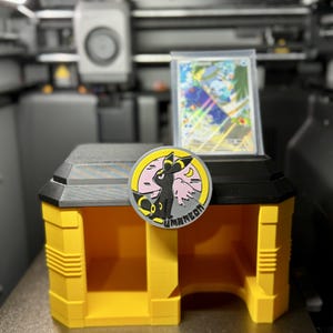 Puede incluir: Un soporte de exhibición impreso en 3D de color amarillo y negro con un emblema circular del personaje Umbreon. El emblema muestra una criatura negra con anillos amarillos sobre un fondo de luna rosa. Una carta coleccionable en funda protectora.