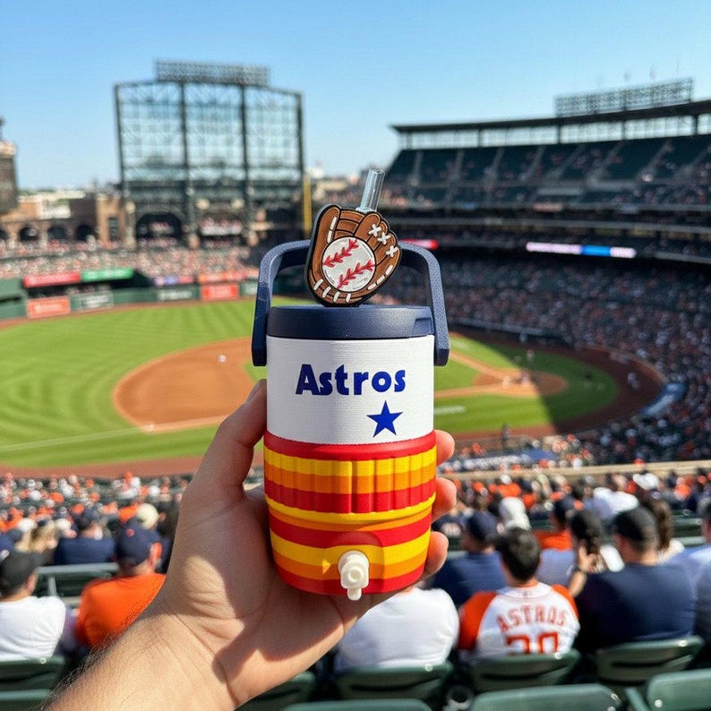 May include: A white and navy blue insulated beverage cooler with a spigot, featuring the word Astros in blue text, a blue star, and horizontal orange and yellow stripes. A baseball glove and ball charm is attached to the top handle with a straw.