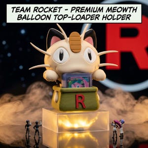 Puede incluir: Un soporte superior para cargador Meowth del Equipo Rocket. El soporte presenta una figura detallada de Meowth con un contenedor verde y la letra "R". La base es un cubo transparente y brillante. El texto "TEAM ROCKET - PREMIUM MEOWTH BALLOON TOP-LOADER HOLDER" está en la parte superior.