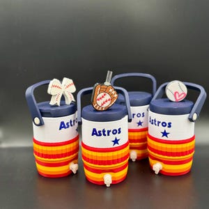 May include: Four miniature cylindrical beverage coolers with navy blue handles and spigots. The bodies are white with the text Astros and a blue star, featuring horizontal orange and red stripes. Toppers include a baseball bow, glove, and ball.