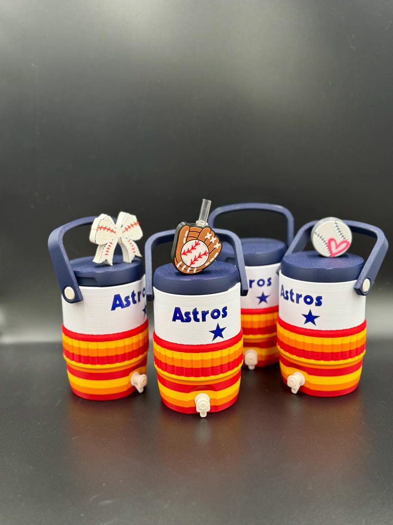 May include: Four miniature cylindrical beverage coolers with navy blue handles and spigots. The bodies are white with the text Astros and a blue star, featuring horizontal orange and red stripes. Toppers include a baseball bow, glove, and ball.