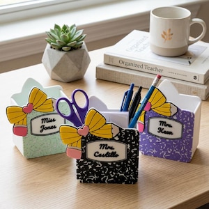 May include: Three desk organizers with pencil and bow accents. Each organizer has a name label: "Miss Jones", "Mrs. Castillo", and "Mrs. Keen". The organizers are filled with pens, pencils, and scissors. A succulent in a geometric pot and a mug are also on the desk.