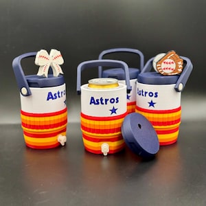 May include: Three cylindrical beverage coolers with navy blue handles and lids, featuring white bodies with the text Astros and a blue star. The lower sections have horizontal red, orange, and yellow stripes. One cooler holds a metal can.