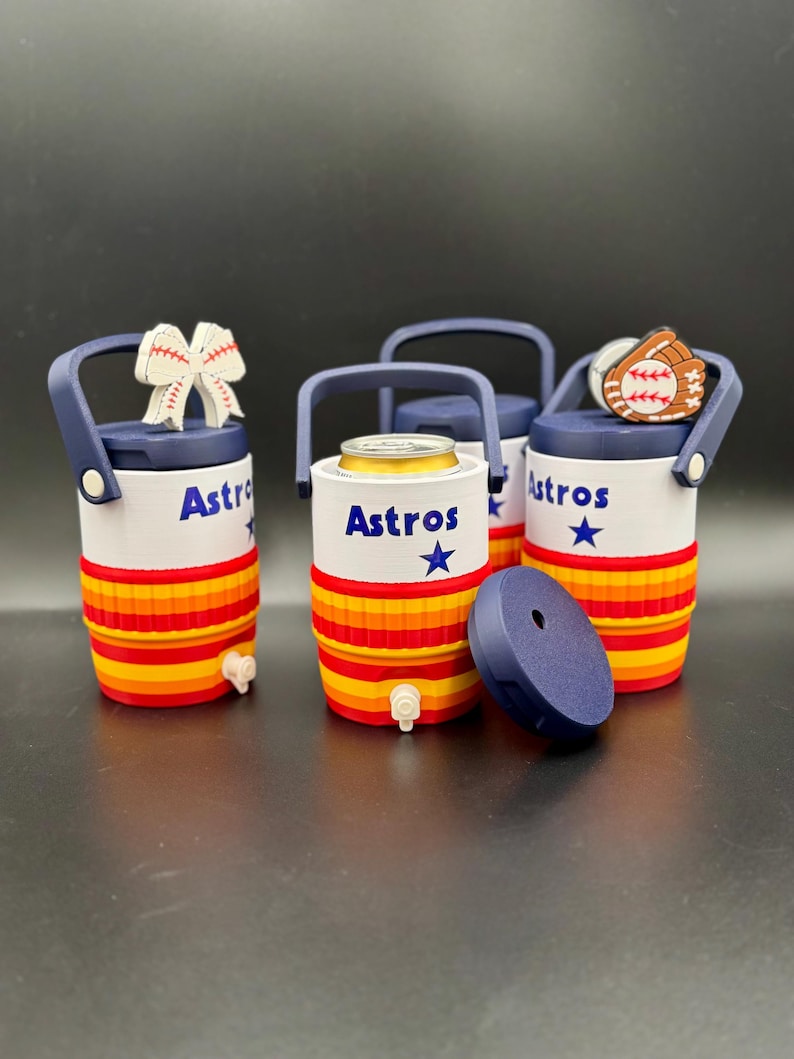 May include: Three cylindrical beverage coolers with navy blue handles and lids, featuring white bodies with the text Astros and a blue star. The lower sections have horizontal red, orange, and yellow stripes. One cooler holds a metal can.