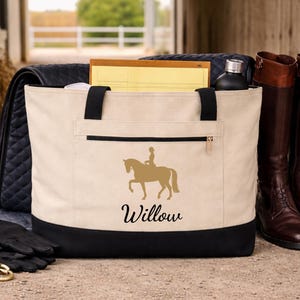 A beige tote bag with a black bottom and handles. A gold silhouette of a horse and rider is on the front, with the name "Willow" below. A pair of brown riding boots stands next to the bag.