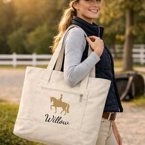 Cream-colored tote bag with a gold equestrian design and the name "Willow" in black script. The bag has a zippered pocket and sturdy handles. The woman in the image is wearing a black cap and vest.