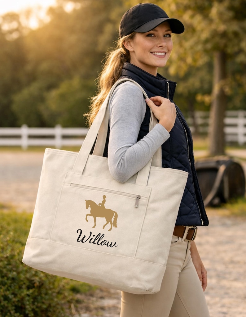 Cream-colored tote bag with a gold equestrian design and the name "Willow" in black script. The bag has a zippered pocket and sturdy handles. The woman in the image is wearing a black cap and vest.