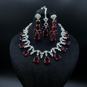 May include: A stunning jewellery set comprising a necklace, earrings, and a headpiece. The set features silver-toned metal with clear and red teardrop-shaped gemstones. The necklace and earrings are displayed on a black mannequin head against a black backdrop.
