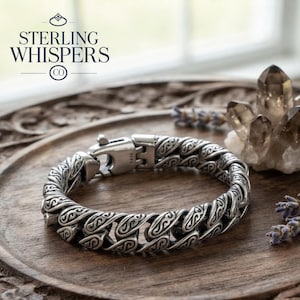 May include: A silver chain bracelet featuring interlocking links with an engraved swirling pattern and a secure lobster claw clasp. The metal surface displays a polished finish with darkened recessed areas to highlight the intricate design details.