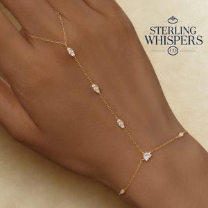 May include: A delicate gold hand chain featuring several clear, oval gemstones. The chain extends from the wrist to the middle finger, with a small heart-shaped gem near the wrist. The text "STERLING WHISPERS CO" is in the upper right corner.