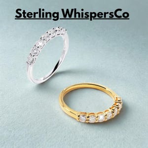May include: Two rings, one silver and one gold, each with a row of clear gemstones. The silver ring is on the top left, and the gold ring is on the bottom right. The rings are set against a light blue background. The text "Sterling WhispersCo" is at the top.