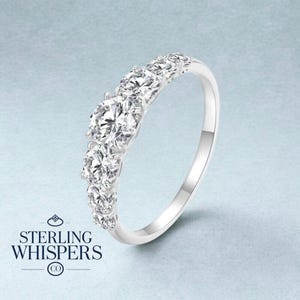 Könnte beinhalten: Ein silberner Ring mit einer Reihe klarer, funkelnder Edelsteine. Der Ring hat ein klassisches Design mit einem Band, das sich um den Finger krümmt. Der Markenname "STERLING WHISPERS" ist unten im Bild zu sehen.