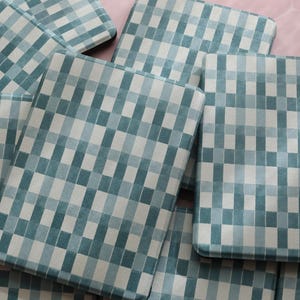 May include: A collection of square, padded items with a blue and white checkered pattern. The items are arranged in a slightly overlapping manner, with a soft, textured appearance. The background is a light pink, hexagonal tile.