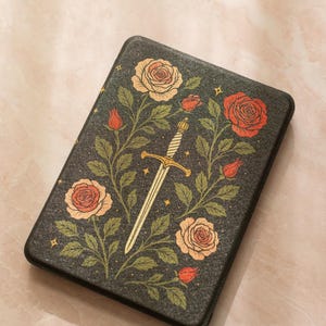 May include: A black rectangular case featuring a floral design with roses in shades of peach and red, green leaves, and a gold sword. The case has a textured appearance and is likely for a tablet or e-reader.