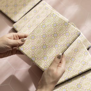 May include: Several square coasters with a repeating geometric pattern in pale yellow, light green, and lavender. The pattern features starbursts and floral elements. The coasters are arranged on a pink surface.