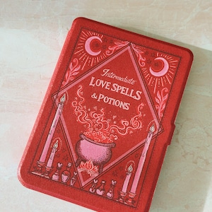 May include: A rectangular red e-reader case featuring a graphic illustration of a cauldron, candles, and small potion bottles. The text "Intermediate LOVE SPELLS & POTIONS" is printed in the center, surrounded by crescent moon and star motifs.