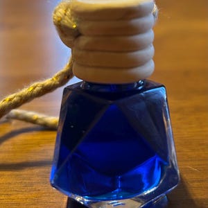 May include: A small, faceted glass bottle filled with vibrant blue liquid. The bottle has a light brown wooden stopper and a gold-colored rope for hanging. The design is geometric and the overall aesthetic is simple and elegant.