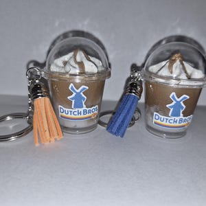 May include: Two miniature coffee cup keychains with clear plastic lids, featuring a brown drink with white and brown swirls, and the Dutch Bros logo. One has an orange tassel, the other a blue tassel.
