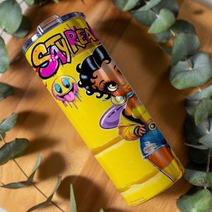 Puede incluir: Un vaso amarillo con una ilustraci&oacute;n de dibujos animados de una mujer y las palabras "Stay Real" en rosa y amarillo. El vaso tambi&eacute;n presenta una carita sonriente con baba rosa goteando. El dise&ntilde;o est&aacute; impreso en un vaso de acero inoxidable.