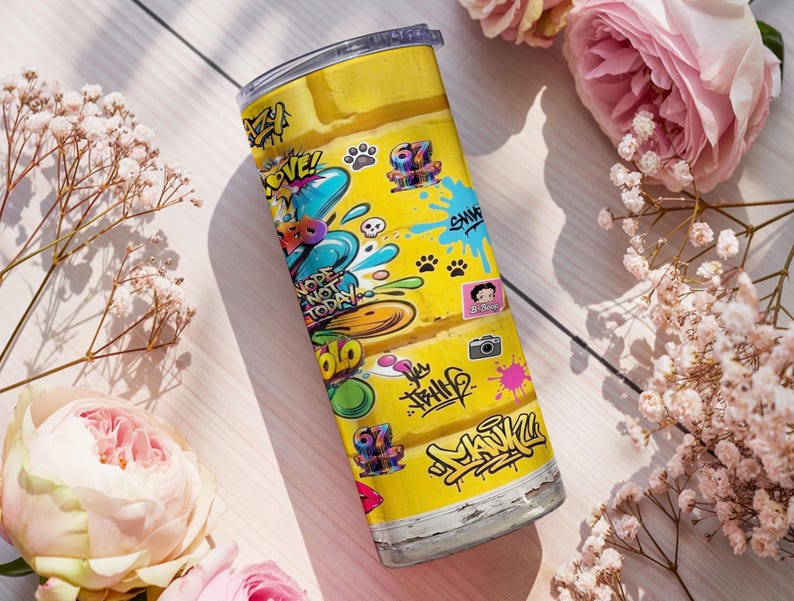 Puede incluir: Un vaso de acero inoxidable amarillo con un dise&ntilde;o de graffiti colorido. El vaso presenta las palabras "Nope Not Today" y otros grafitis. El dise&ntilde;o incluye personajes de dibujos animados, huellas de patas y un icono de c&aacute;mara.
