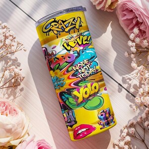 Puede incluir: Un vaso de acero inoxidable amarillo con un dise&ntilde;o de estilo grafiti. El vaso presenta las palabras "Crazy", "Love", "Nope Not Today" y "Yolo" en varios colores. Tambi&eacute;n presenta un par de labios rojos.