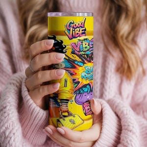 Puede incluir: Un vaso amarillo con un dise&ntilde;o colorido de "Good Vibe". El vaso presenta un dise&ntilde;o vibrante de estilo graffiti con las palabras "Good Vibe" y otro texto. El vaso es sostenido por una persona que lleva un su&eacute;ter rosa.