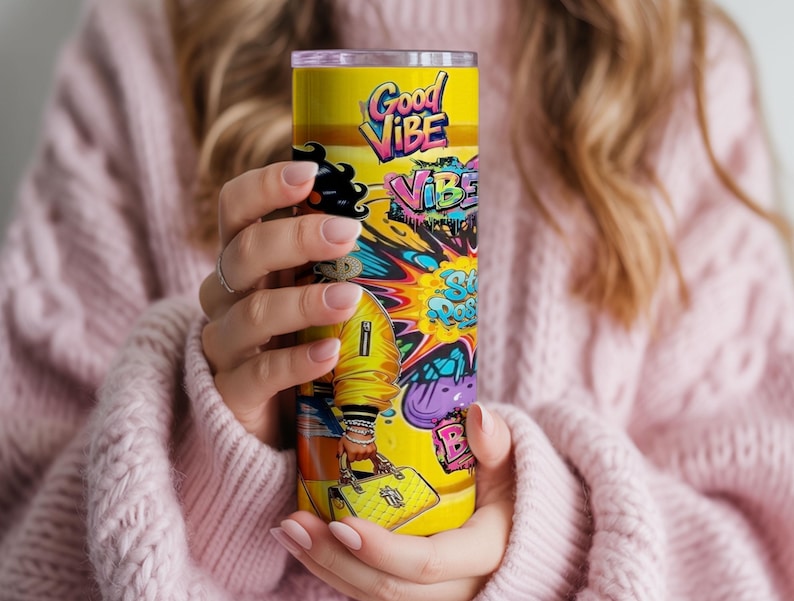 Puede incluir: Un vaso amarillo con un dise&ntilde;o colorido de "Good Vibe". El vaso presenta un dise&ntilde;o vibrante de estilo graffiti con las palabras "Good Vibe" y otro texto. El vaso es sostenido por una persona que lleva un su&eacute;ter rosa.