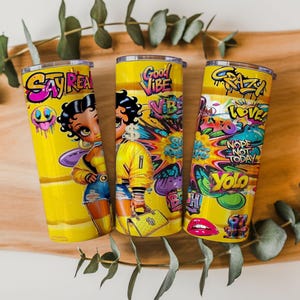 Puede incluir: Tres vasos t&eacute;rmicos amarillos con gr&aacute;ficos de dibujos animados coloridos y texto. Uno presenta un personaje de dibujos animados y las palabras "Say Rea". Otro tiene "Good Vibe" y una figura de dibujos animados. El tercero tiene "Crazy", "Love", "Nope Not Today" y "Yolo".
