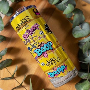 Puede incluir: Un vaso de acero inoxidable amarillo con un dise&ntilde;o de estilo grafiti. El vaso presenta la palabra "Boop" en letras grandes y coloridas, junto con otros grafitis, calaveras y el texto "B. Boop". El vaso es cil&iacute;ndrico y tiene una tapa plateada.