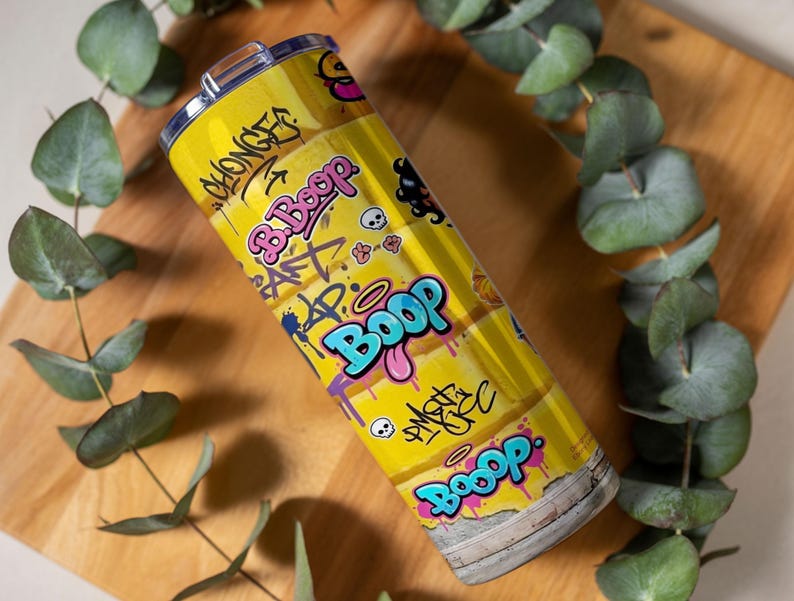 Puede incluir: Un vaso de acero inoxidable amarillo con un dise&ntilde;o de estilo grafiti. El vaso presenta la palabra "Boop" en letras grandes y coloridas, junto con otros grafitis, calaveras y el texto "B. Boop". El vaso es cil&iacute;ndrico y tiene una tapa plateada.