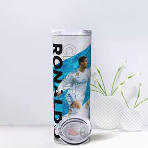 K&ouml;nnte beinhalten: Ein wei&szlig;-blauer Becher mit einem Fu&szlig;ballspieler in wei&szlig;er Uniform. Der Name "CRISTIANO RONALDO" ist vertikal in Schwarz und Wei&szlig; aufgedruckt. Der Becher hat einen transparenten Deckel und steht vor einem wei&szlig;en Hintergrund.