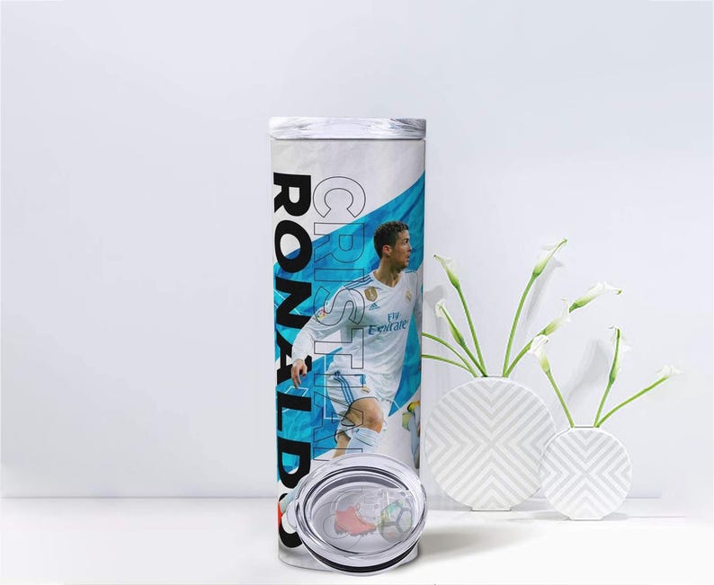 K&ouml;nnte beinhalten: Ein wei&szlig;-blauer Becher mit einem Fu&szlig;ballspieler in wei&szlig;er Uniform. Der Name "CRISTIANO RONALDO" ist vertikal in Schwarz und Wei&szlig; aufgedruckt. Der Becher hat einen transparenten Deckel und steht vor einem wei&szlig;en Hintergrund.