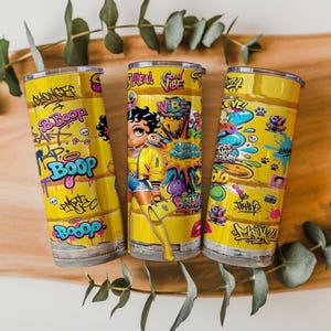 Puede incluir: Tres vasos aislados amarillos con arte de graffiti y un personaje de dibujos animados. Los vasos presentan las palabras "Boop" y "Good Vibe" en un estilo graffiti. El personaje de dibujos animados lleva una chaqueta amarilla y sostiene una bolsa morada.
