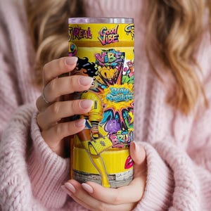Puede incluir: Un vaso amarillo con un dise&ntilde;o vibrante con las palabras "Good Vibe" y "Stay Positive". El vaso muestra una ilustraci&oacute;n de dibujos animados de una mujer con un atuendo amarillo, con coloridos acentos de estilo graffiti. El vaso es sostenido por una persona que lleva un su&eacute;ter rosa.