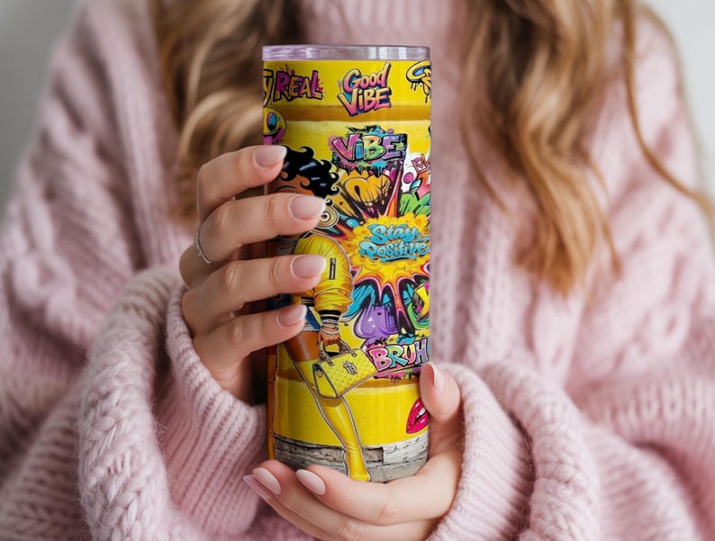 Puede incluir: Un vaso amarillo con un dise&ntilde;o vibrante con las palabras "Good Vibe" y "Stay Positive". El vaso muestra una ilustraci&oacute;n de dibujos animados de una mujer con un atuendo amarillo, con coloridos acentos de estilo graffiti. El vaso es sostenido por una persona que lleva un su&eacute;ter rosa.