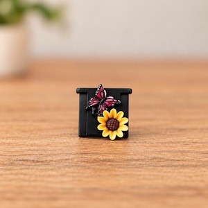 May include: A square black button featuring a raised, detailed pink and black butterfly positioned above a bright yellow sunflower with a brown center. The design is centered on the flat black surface of the button, which has a smooth finish.