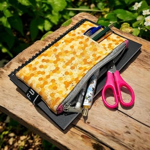 May include: A yellow and orange patterned pencil case with a silver zipper, containing pens and pencils, rests on a black notebook. A pair of pink scissors is next to the pencil case. The items are on a wooden surface.