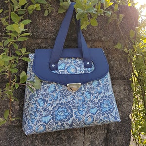 May include: A blue and white floral tote bag with a navy blue flap and a gold clasp. The bag is 13 inches wide, 11 inches high, and 5 inches deep. The bag is sitting on a stone wall with green vines.