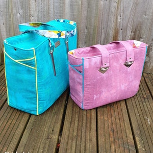 Puede incluir: Dos bolsas de mano, una turquesa con ribete amarillo y otra rosa con ribete azul. Las bolsas están sobre una plataforma de madera. El texto "The Piped Pocket Tote" está en la parte superior de la imagen. El texto "Finished sizes approx:" está debajo de las bolsas. El texto "Large: 13 1/2" x 11 1/2" x 5 1/2" (35cm x 29cm x 14cm)" está debajo del texto "Finished sizes approx:". El texto "Small: 11 1/2" x 9 1/2" x 4 1/2" (29cm x 23.5cm x 11.5cm)" está debajo del texto "Large: 13 1/2" x 11 1/2" x 5 1/2" (35cm x 29cm x 14cm)". El texto "SEWING PATTERNS by Mrs h." está en la esquina inferior derecha de la imagen.