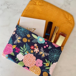 May include: A floral print clutch with a navy blue background, featuring pink, orange, and teal flowers and butterflies. The interior is mustard yellow and holds cosmetics and a white card with "No 7" printed on it.